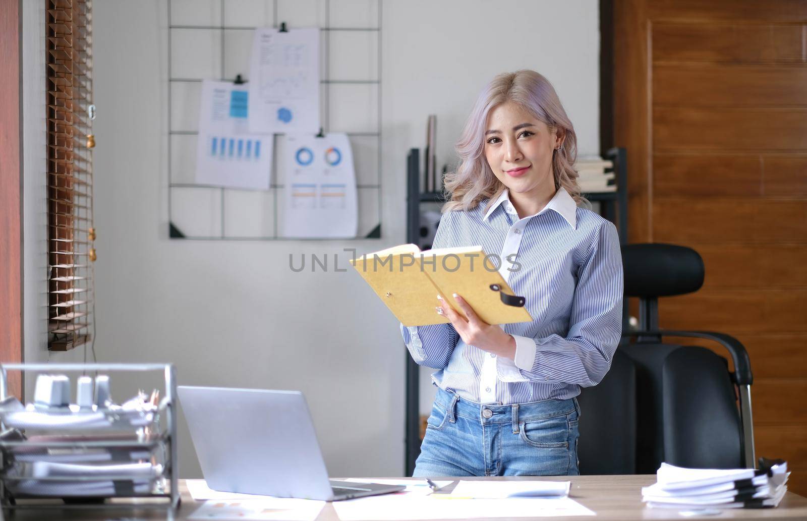 Charming young Asian businesswoman with a smile standing holding book at the office. Looking at camera..
