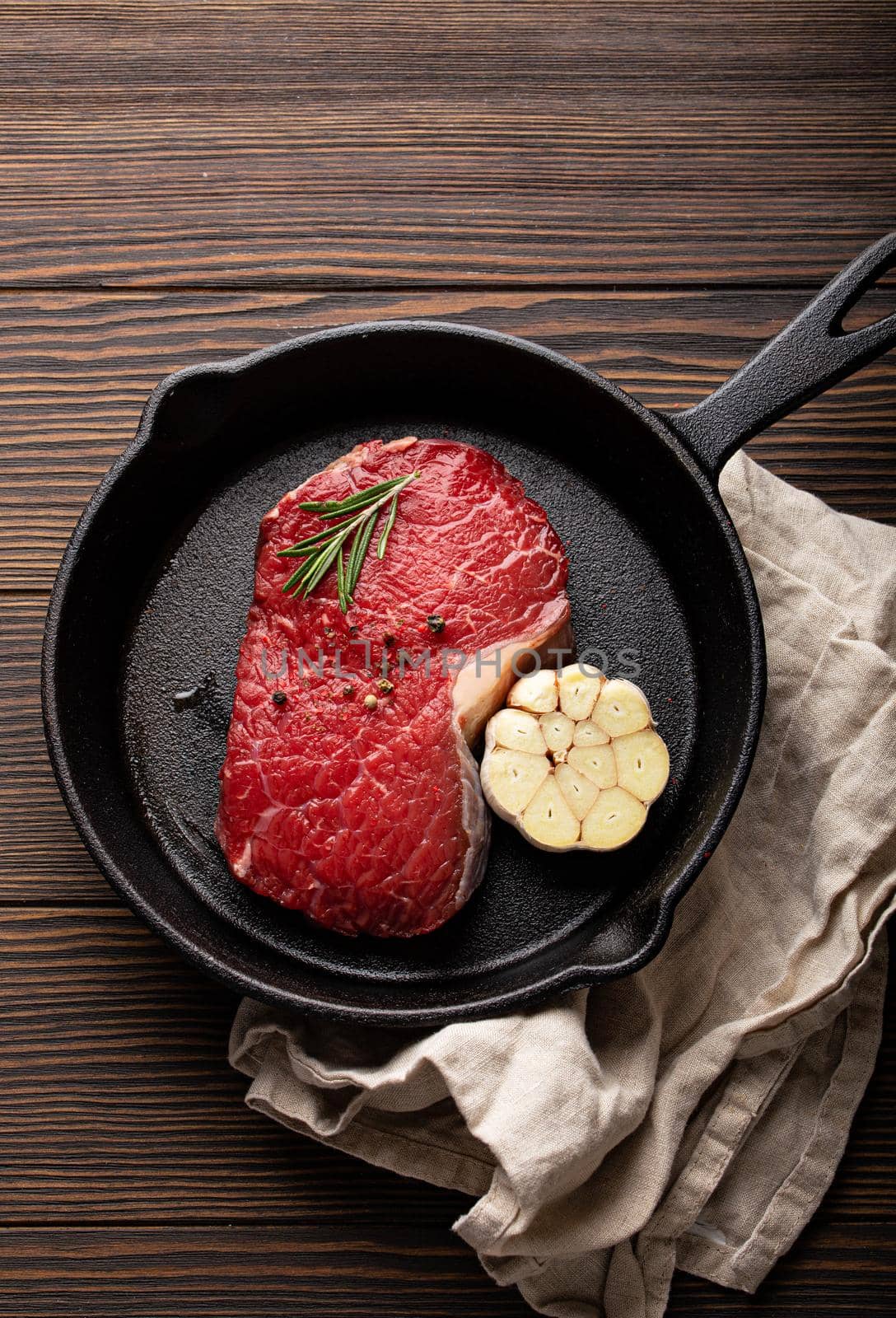 Raw fresh beef steak with seasonings, garlic and rosemary on cast iron frying pan on dark wooden background from above with textile napkin
