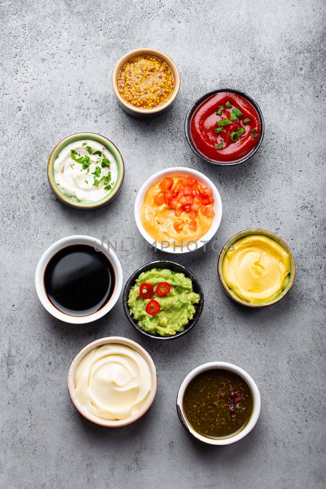 Set of different sauces in bowls on gray rustic concrete background, top view, close-up. Tomato ketchup, mayonnaise, guacamole, mustard, soy sauce, pesto, cheese sauce - assortment of dips