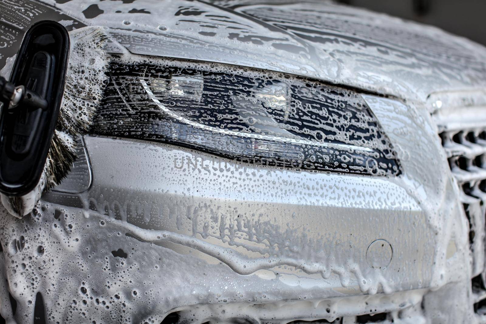 Front light of silver car washed in manual carwash, brush leaving strokes in shampoo and foam.