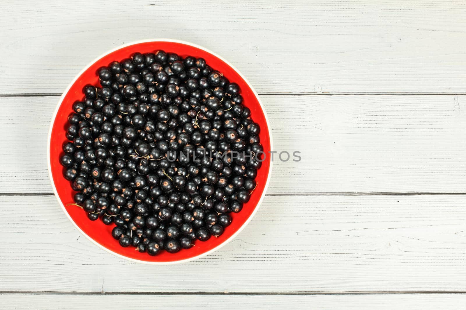 Tabletop view, red ceramic bowl full of freshly harvested blackcurrants, on white boards desk.