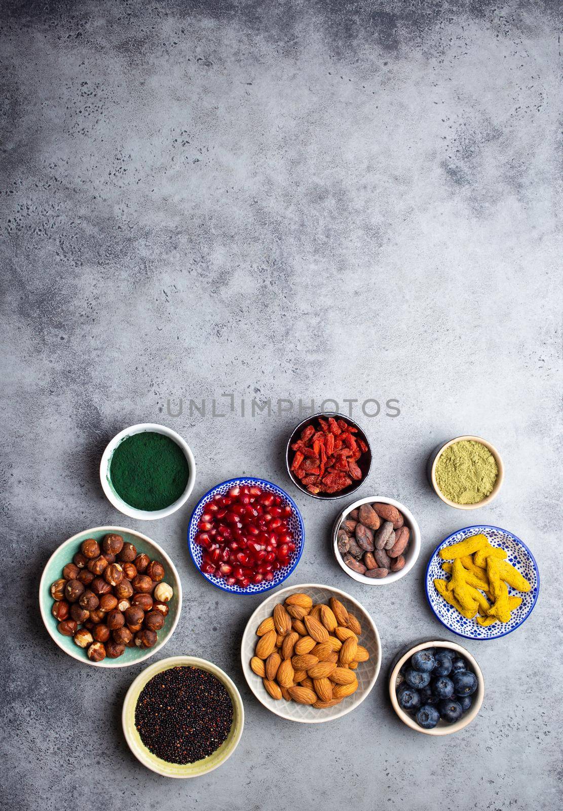 Set of different superfoods in bowls on stone gray background: spirulina, goji berry, cocoa, matcha green tea, quinoa, chia seeds, blueberries, nuts for happy healthy living, top view, copy space