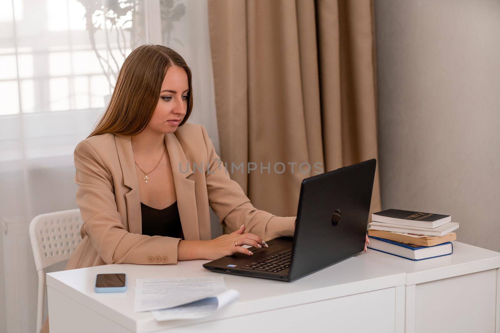 European professional woman sitting with laptop at home office desk, positive woman studying while working on PC. She is wearing a beige jacket and jeans. by Matiunina