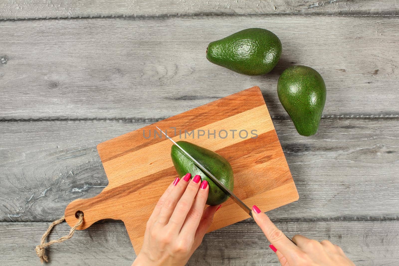 Tabletop view, young woman cutting whole avocado on chopping board.