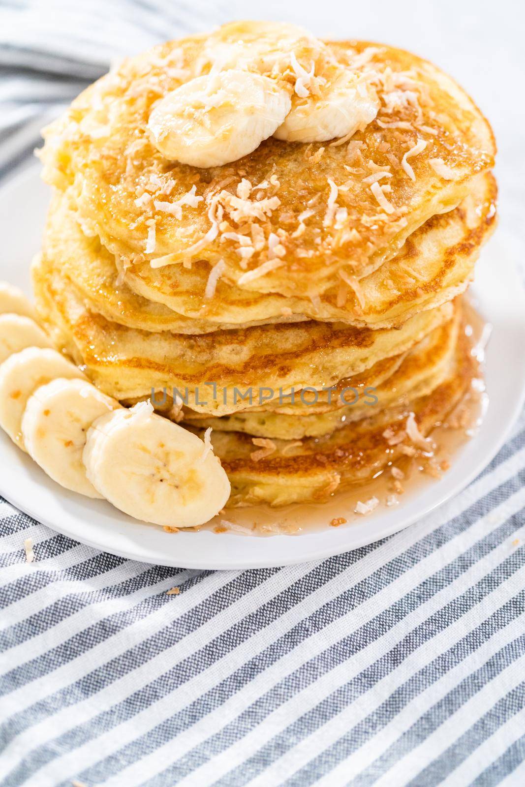 Stack of freshly baked coconut banana pancakes garnished with sliced bananas, toasted coconut, and maple syrup.