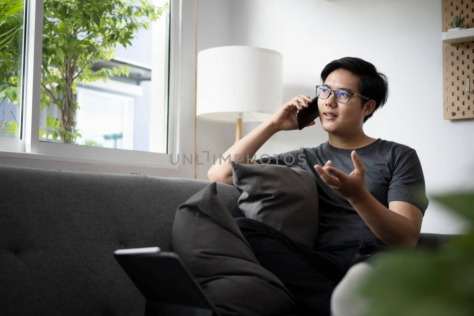 Young Asian man sitting on couch at home and talking on mobile phone. by prathanchorruangsak