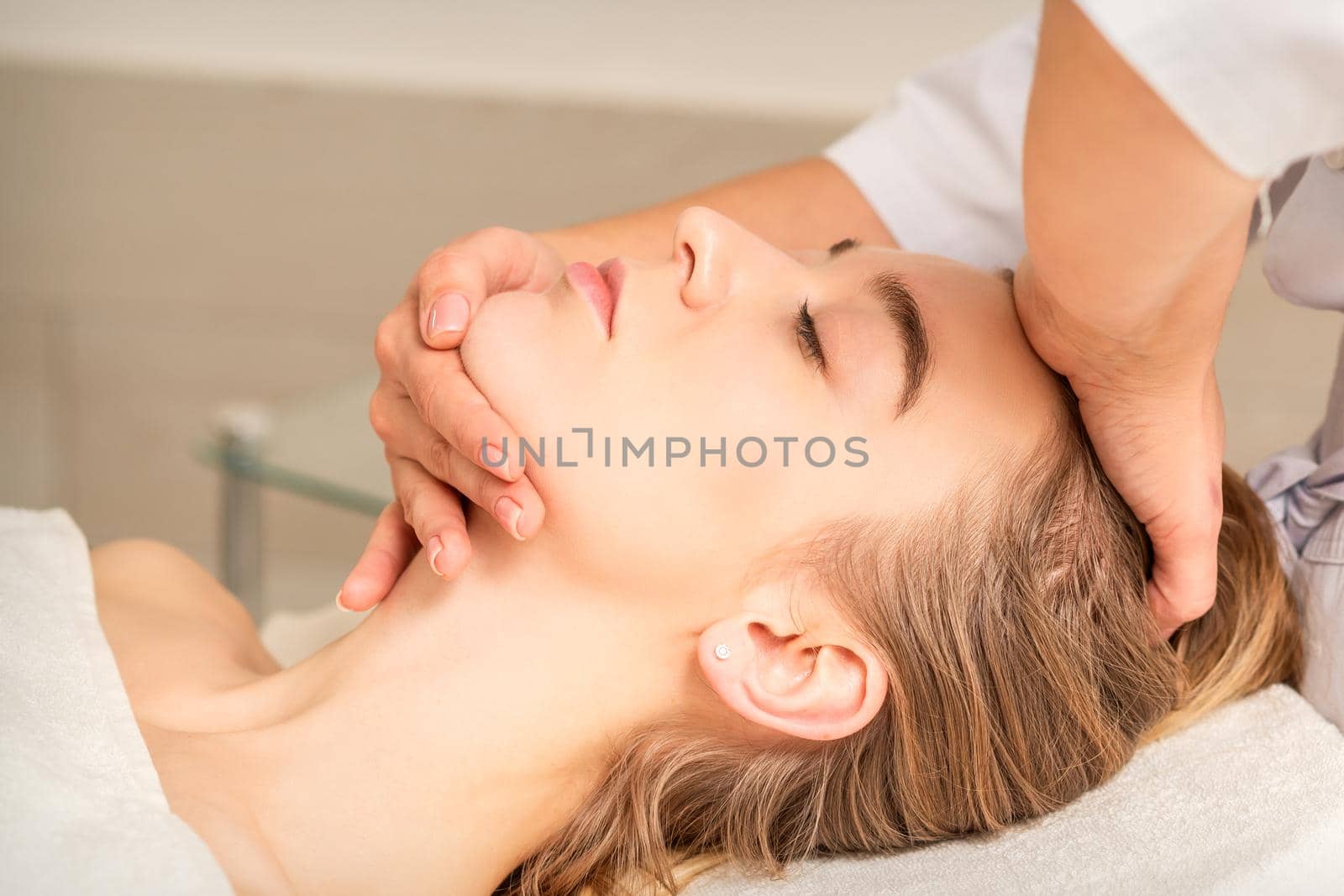 Cosmetologist hands doing facial massage on forehead and chin of young female at spa salon