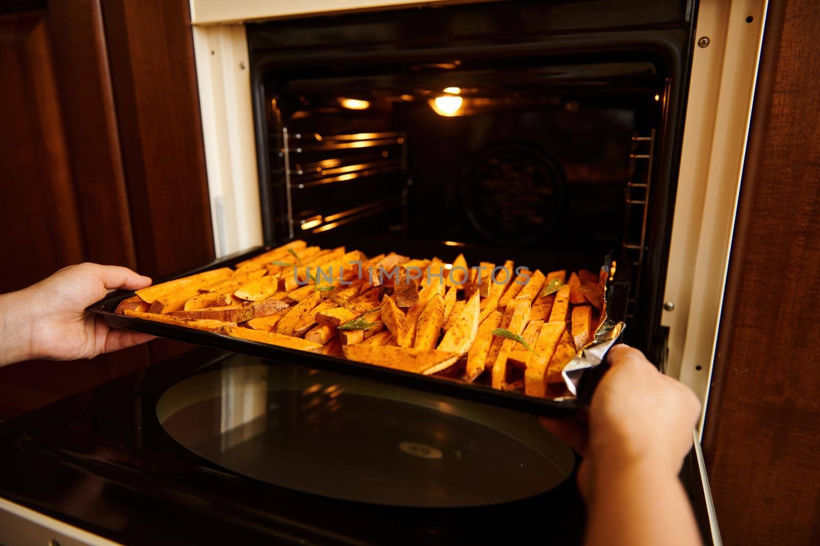 Close-up of housewife's hands putting a baking sheet with sweet potatoes, seasoned with spices and herbs into an oven by artgf