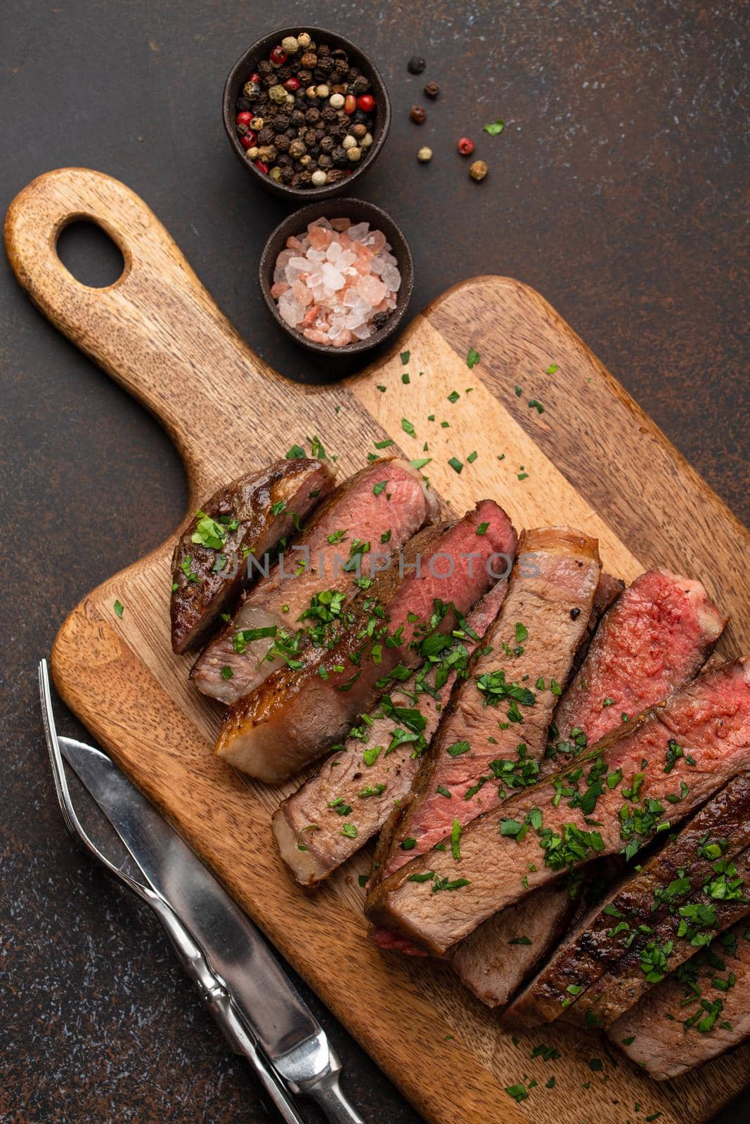 Grilled or fried prime marbled meat steak Ribeye sliced and served on wooden cutting board with cutlery. Top view of juicy cooked beef steak cut in slices on rustic brown concrete background