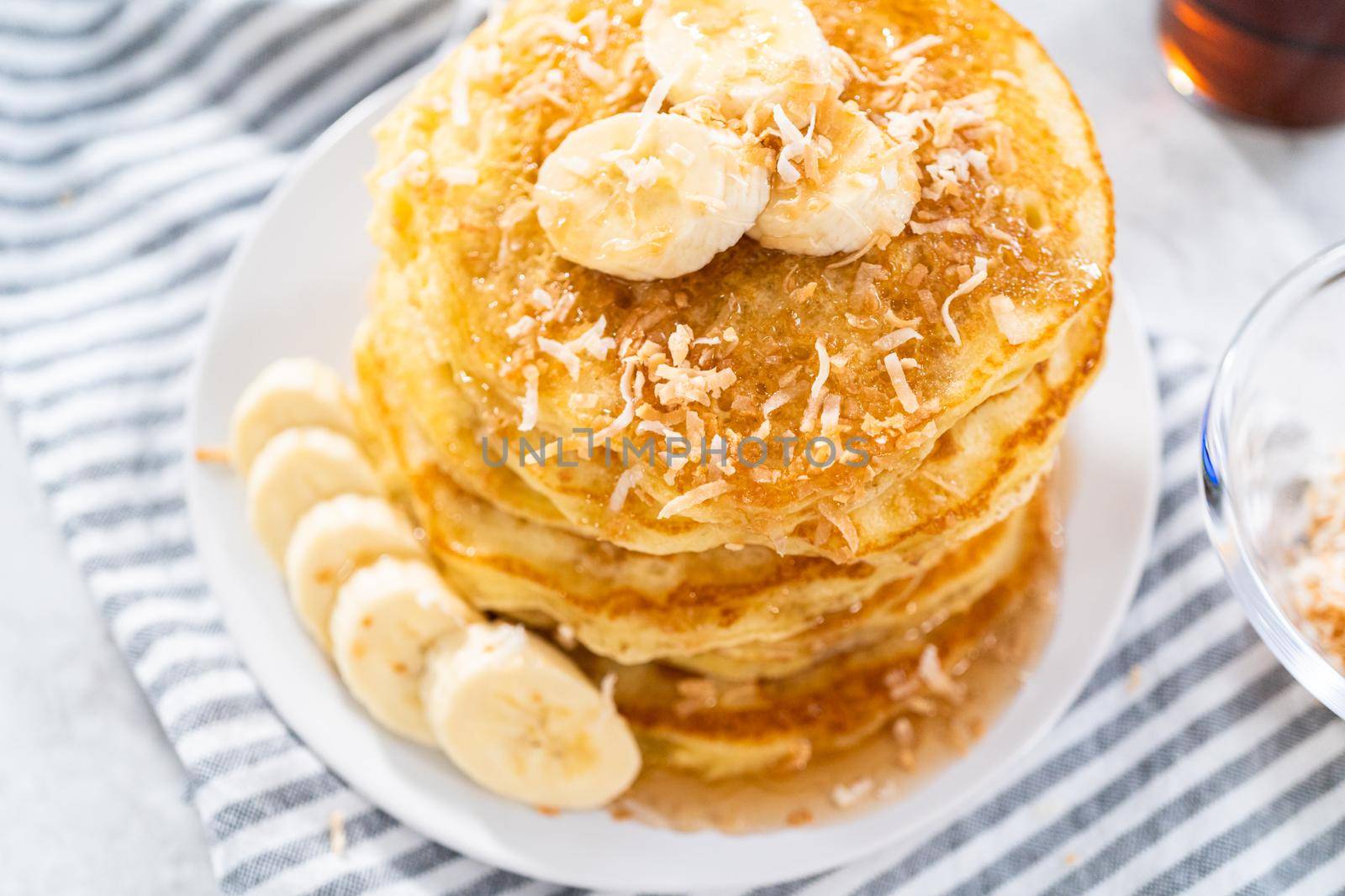 Stack of freshly baked coconut banana pancakes garnished with sliced bananas, toasted coconut, and maple syrup.