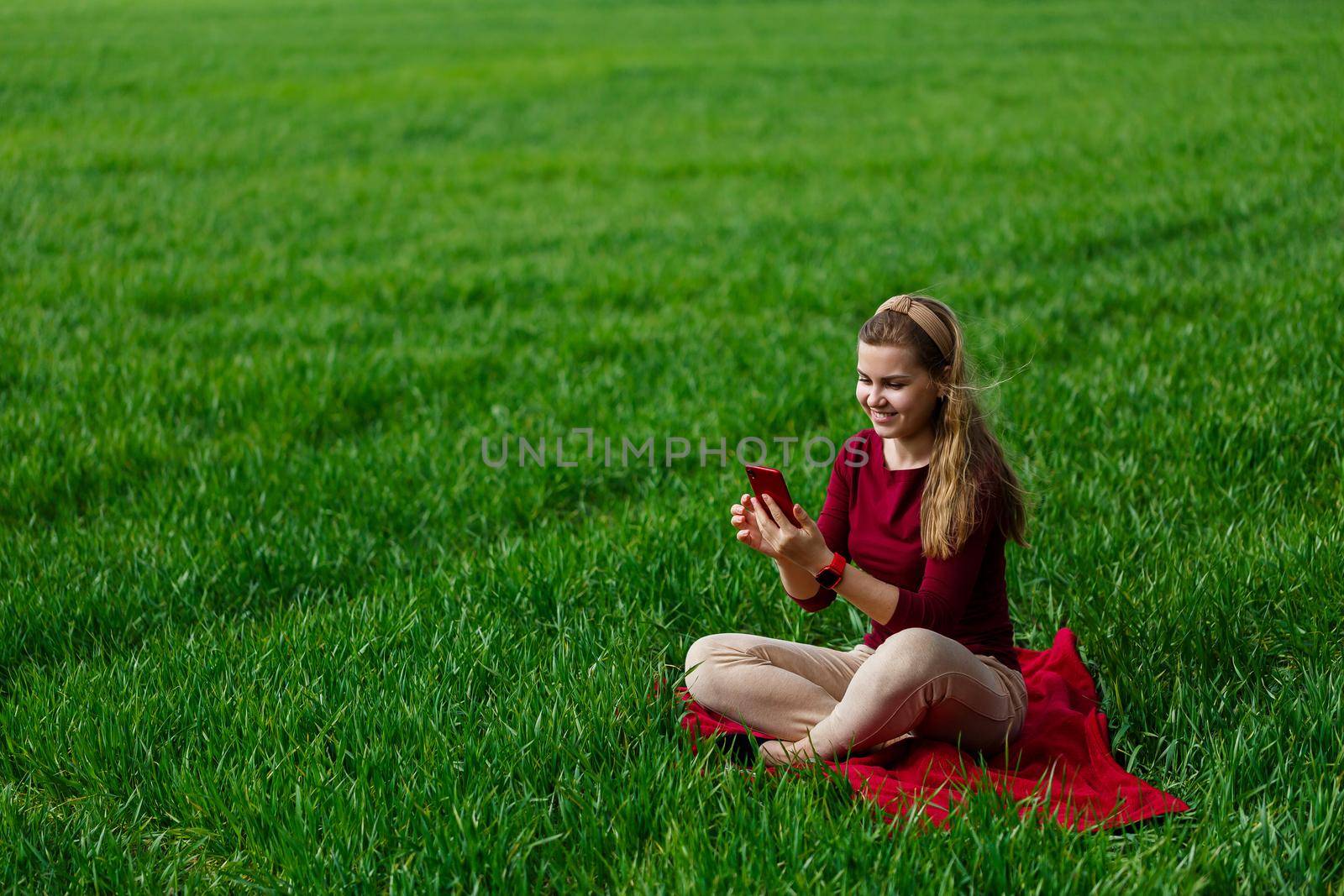 Beautiful woman with a phone in her hands sits on the grass. The girl looks at herself in the camera of the phone and takes selfie photos. She smiles and enjoys a warm day. Concept photo on smartphone