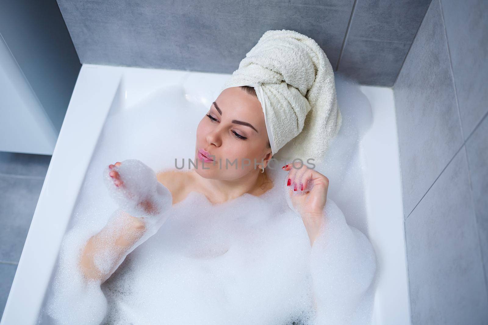 Girl woman with a white towel on her head lies in the bathroom in a white bath. In the hands of a lot of muddy soap. Full whirlpool foam. Relax after a hard day. Spa relaxing treatment