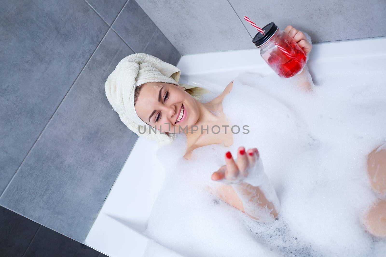 Beautiful young woman with a towel on her head, drinking a cocktail, takes a bath at home. Relax after a hard day.