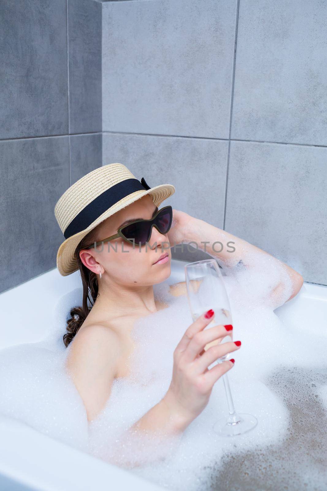 Beautiful young woman in a hat on her head and sunglasses drinking a cocktail takes a bath at home. Relax after a hard day. The spa is a relaxing procedure.
