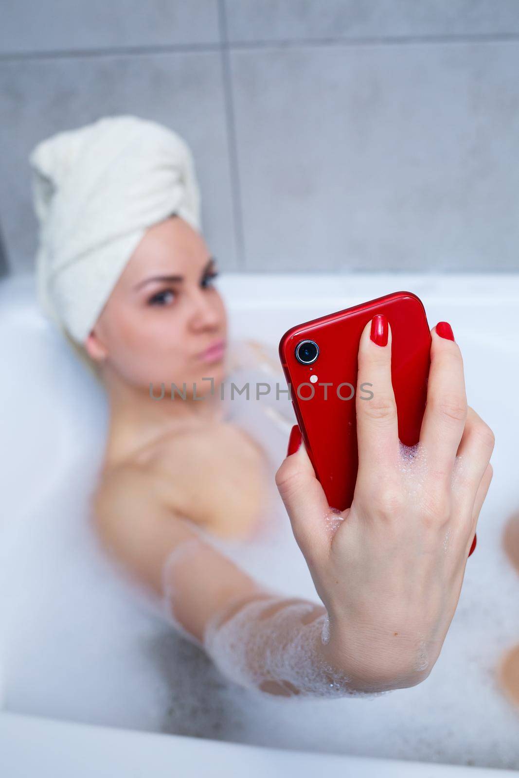 Girl woman with a towel on her head lies with a glass of champagne in a white bathtub and takes a selfie on the phone. In her hands a lot of drunk soap. Relax after a hard day. Spa relaxing procedure