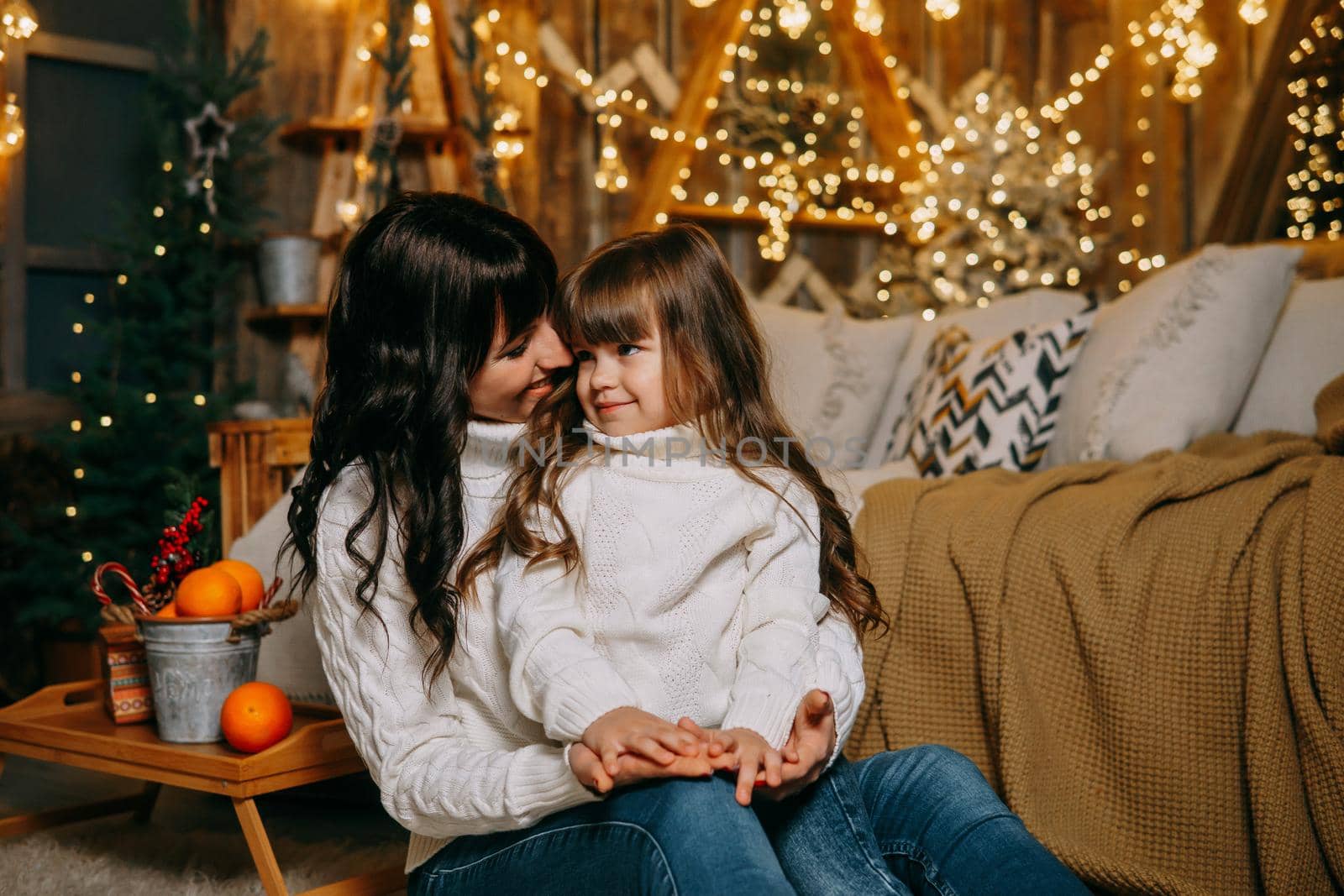 A little girl with her mother in a cozy home environment on the sofa next to the Christmas tree. The theme of New Year holidays and festive interior with garlands and light bulbs