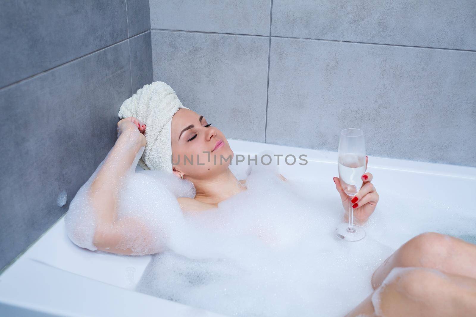 Young beautiful woman with a white towel on her head lies in a white bath. In the hands of a glass of white wine. Relax after a hard day. Relaxing spa treatments
