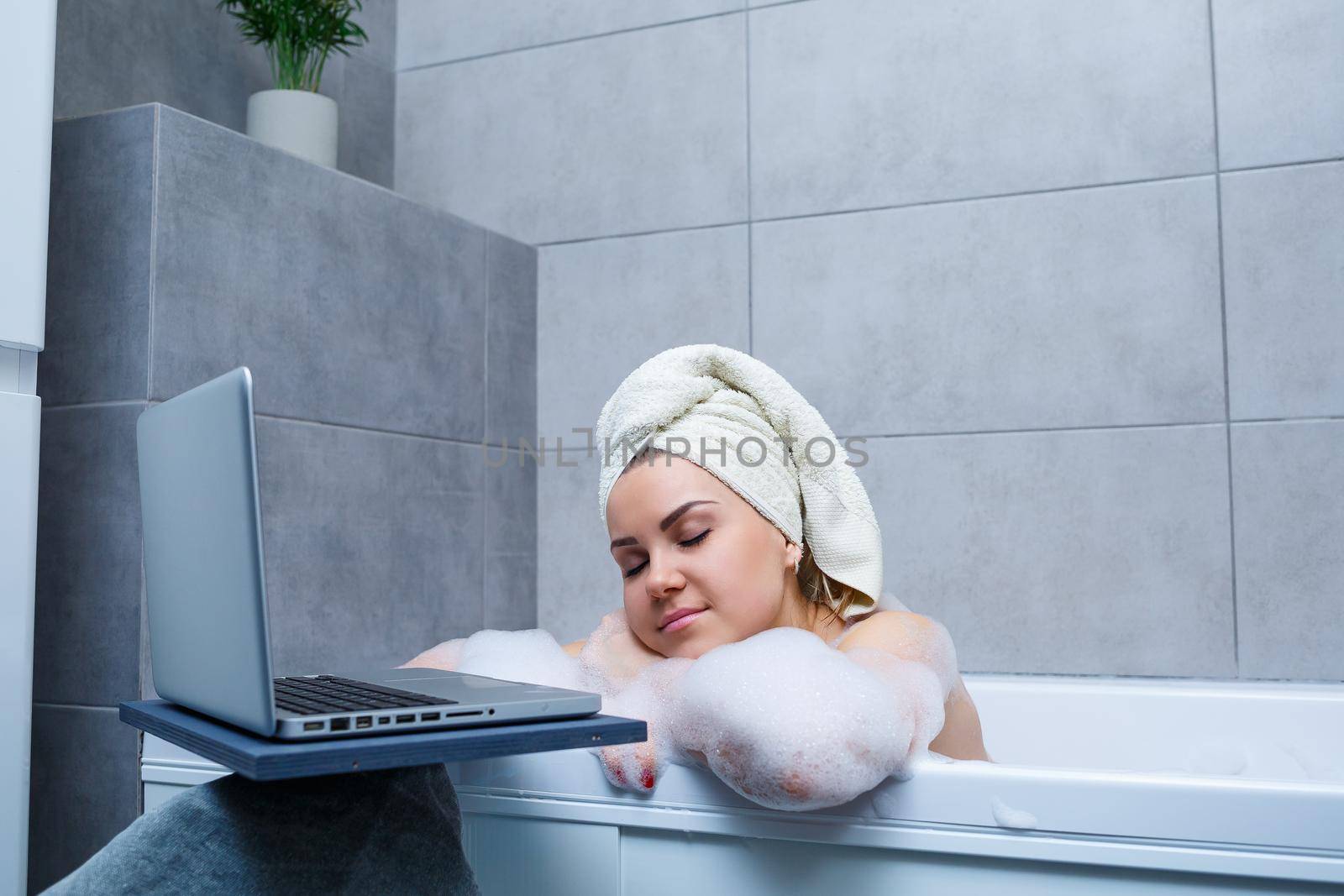 A young woman with a towel on her head is watching a movie on a laptop while sitting in a bath in a beauty salon. Relax in the bathroom without linen. Body care and relaxation concept.