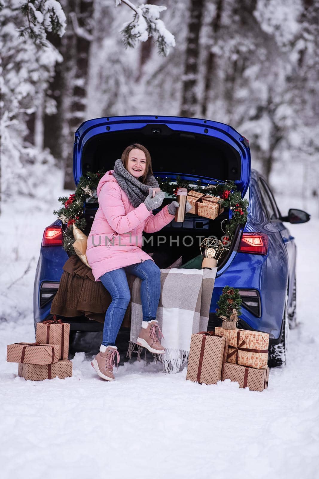 A woman in winter clothes drinks a hot drink sitting in a car decorated in a New Year's way. Traveling by car through the forest. A trip before Christmas.