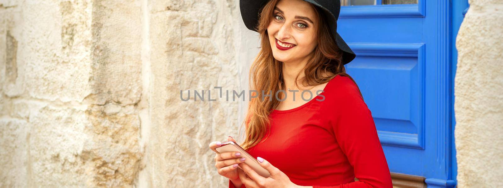 Portrait of tourist young caucasian woman in a red long dress and black hat with suitcase sitting on the stairs outdoors