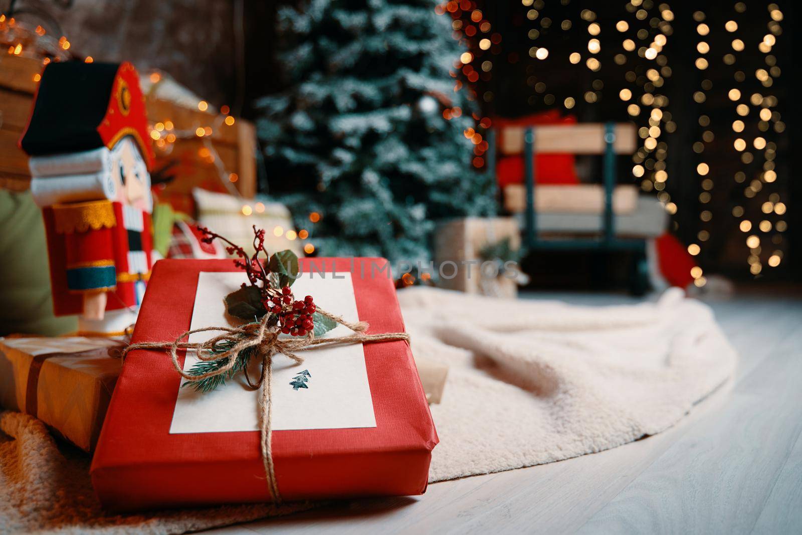 Close-up of gift box and New Year's atmosphere on background. Christmas tree, plaid with colorful pillows on floor, Nutcracker figurine and bright garlands on walls. Festive interior in photo studio.
