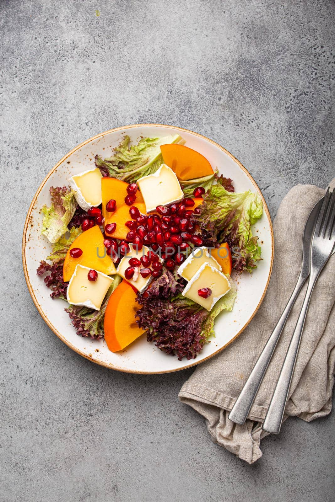 Top view flat lay persimmon salad with brie cheese, fresh salad leaves, pomegranate seeds on white plate and stone gray background, seasonal fruit salad as appetizer, vegetarian healthy food