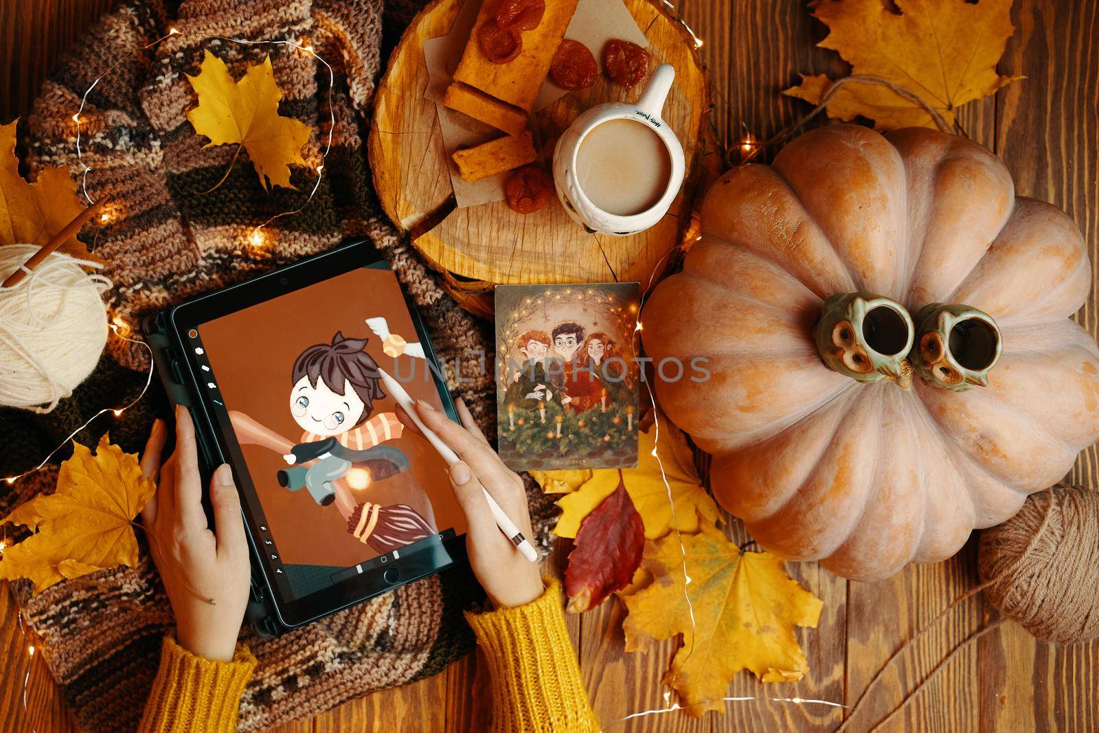 Scarf, coffee, pie, pumpkin, leaves and garland on a wooden table. Young woman in a sweater draws on a tablet. Harry Potter plays Quidditch on a broomstick. Bishkek, Kyrgyzstan - November 1, 2019.