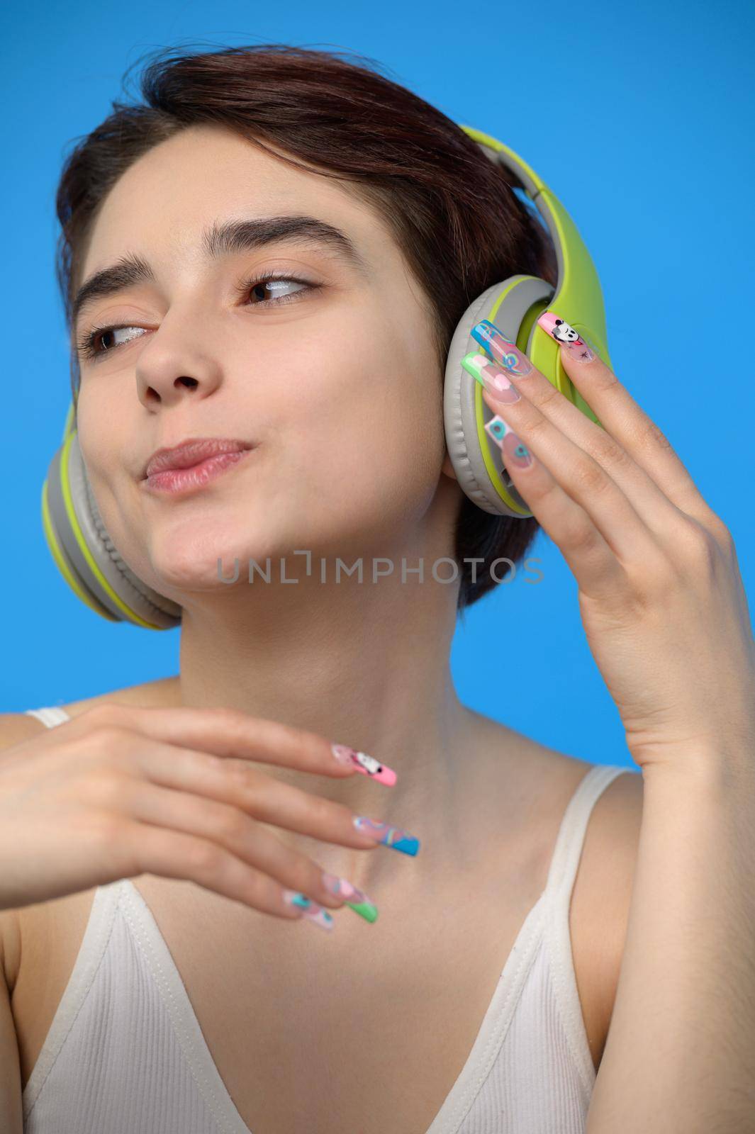 Portrait of cute young bare shoulders brunette with extravagant nail art listening to music using wireless headphones, studio shot