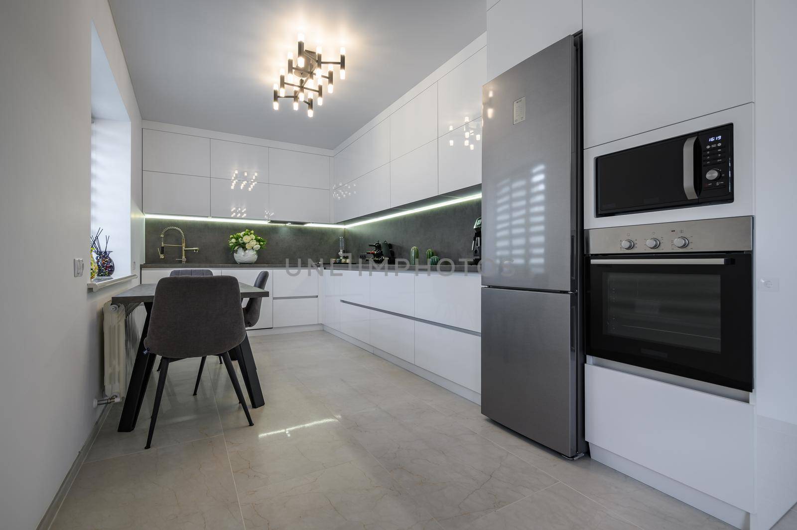 Luxurious modern trendy white and grey kitchen interior after renovation, with granite counter top and marble floor, dining table next to window