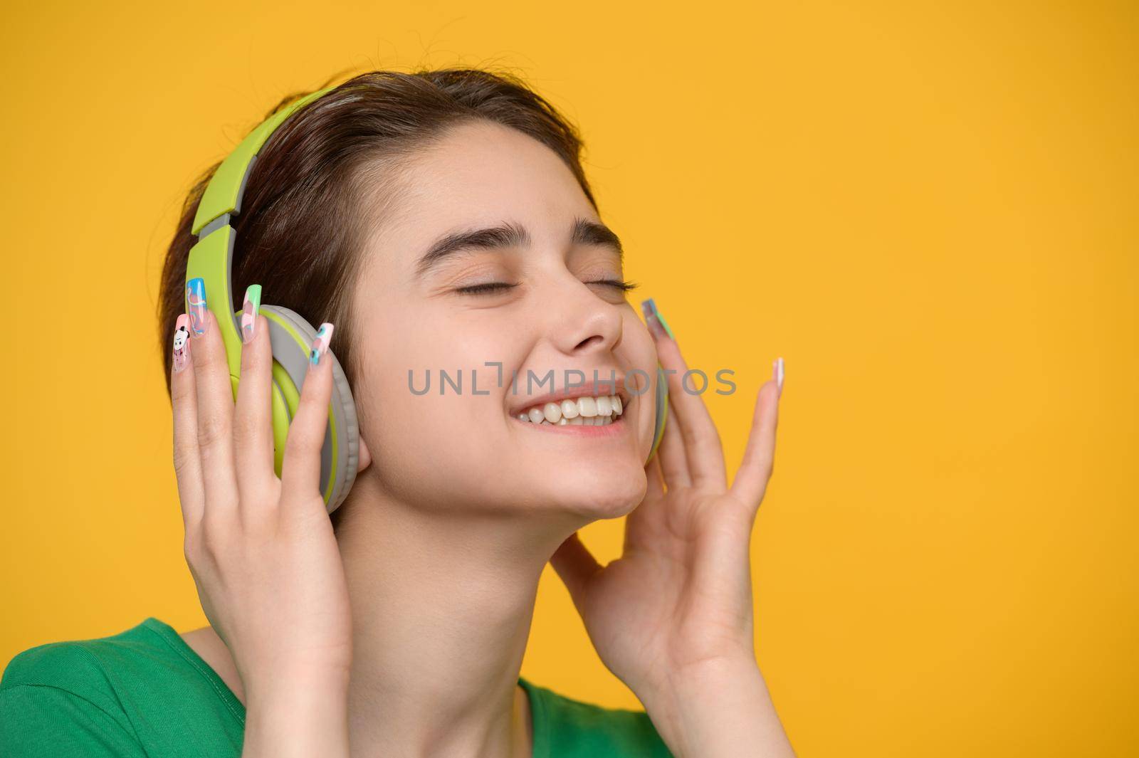 Young beautiful brunette with a short haircut enjoys listening to music on wireless headphones, studio shot at yellow packground