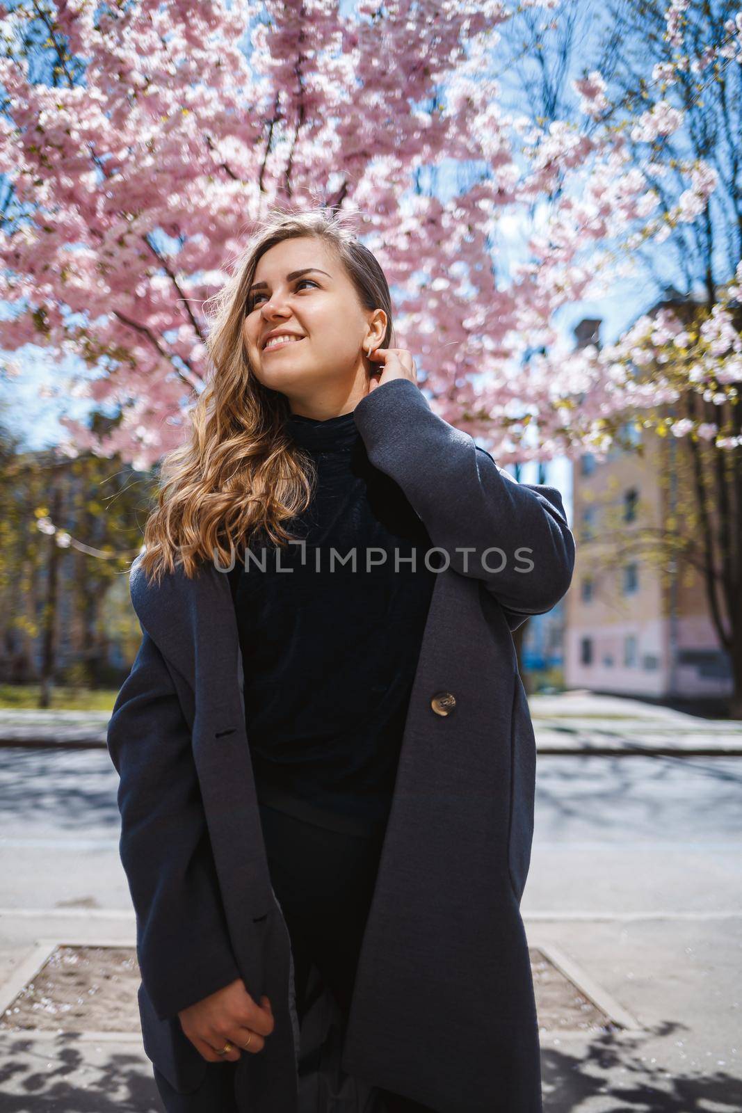 A young slender female model with long wavy hair and, dressed in a gray coat, sneakers, stands on the street near a flowering shrub with beautiful pink flowers in the background and poses.
