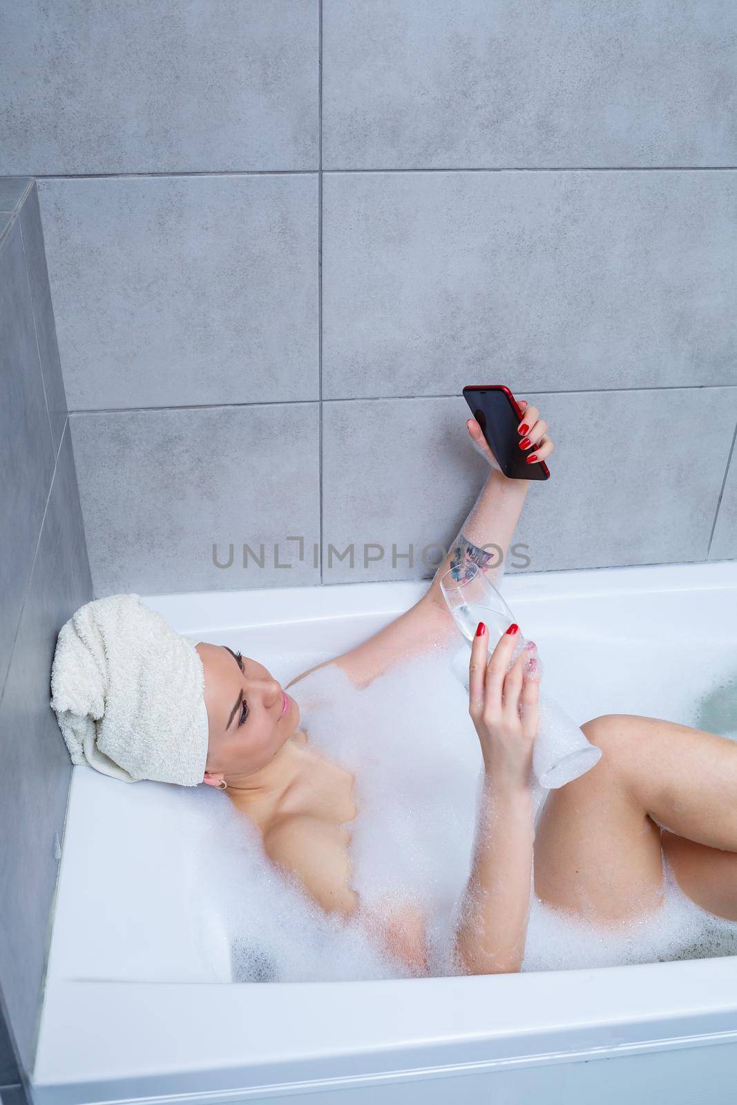 Girl woman with a towel on her head lies with a glass of champagne in a white bathtub and takes a selfie on the phone. In her hands a lot of drunk soap. Relax after a hard day. Spa relaxing procedure