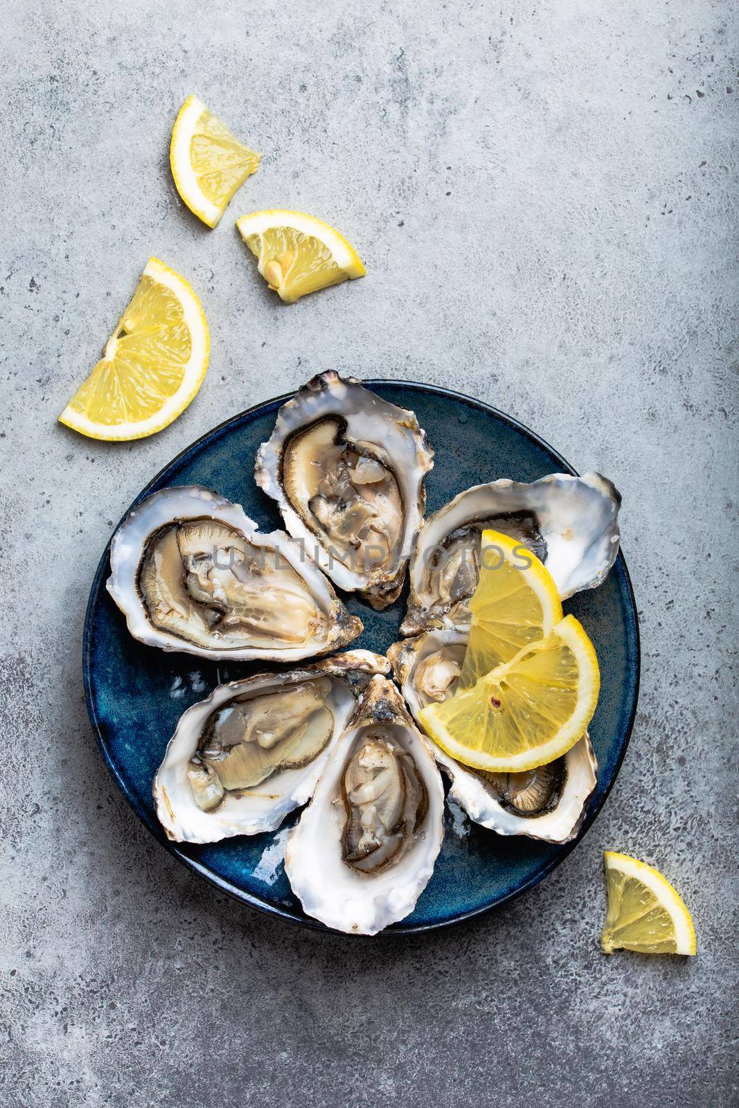 Set of half dozen fresh opened oysters in shell with lemon wedges served on rustic blue plate on gray stone background, close up, top view