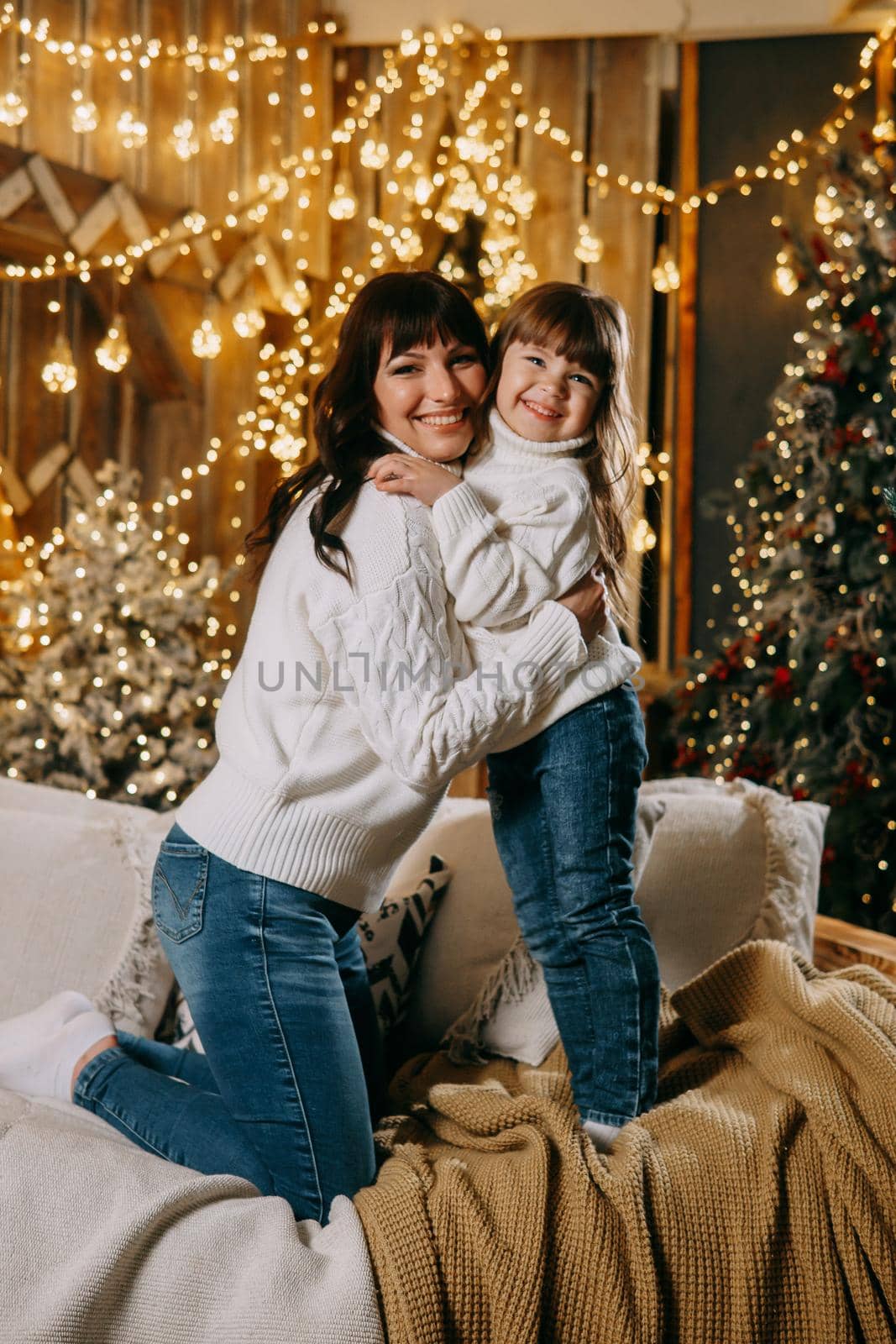 A little girl with her mother in a cozy home environment on the sofa next to the Christmas tree. The theme of New Year holidays and festive interior with garlands and light bulbs