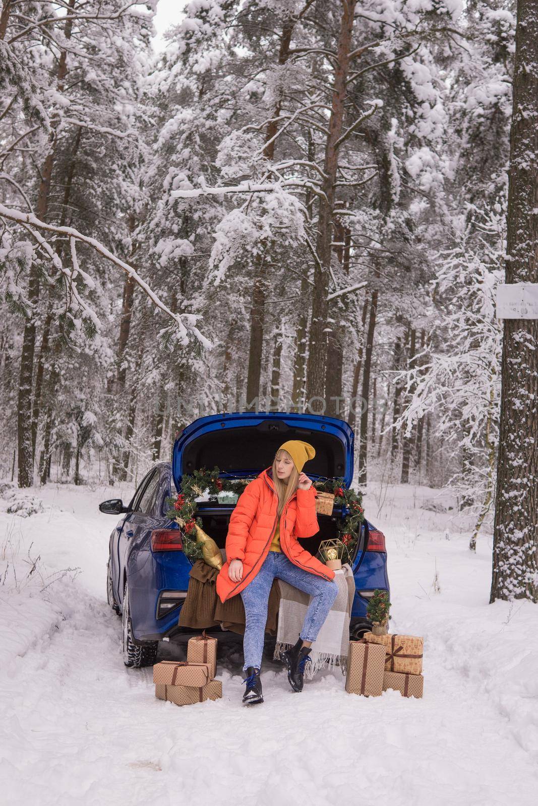 A woman in a winter snow-covered forest in the trunk of a blue car decorated with Christmas decor. The concept of Christmas and winter holidays by Annu1tochka