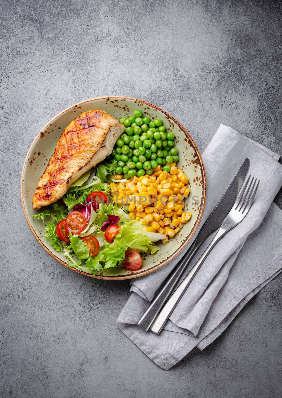 Baked chicken breast on plate with fresh salad, green peas and corn, grey stone background, top view. Healthy fitness meal with chicken fillet, balanced in proteins and carbs. Dieting and nutrition
