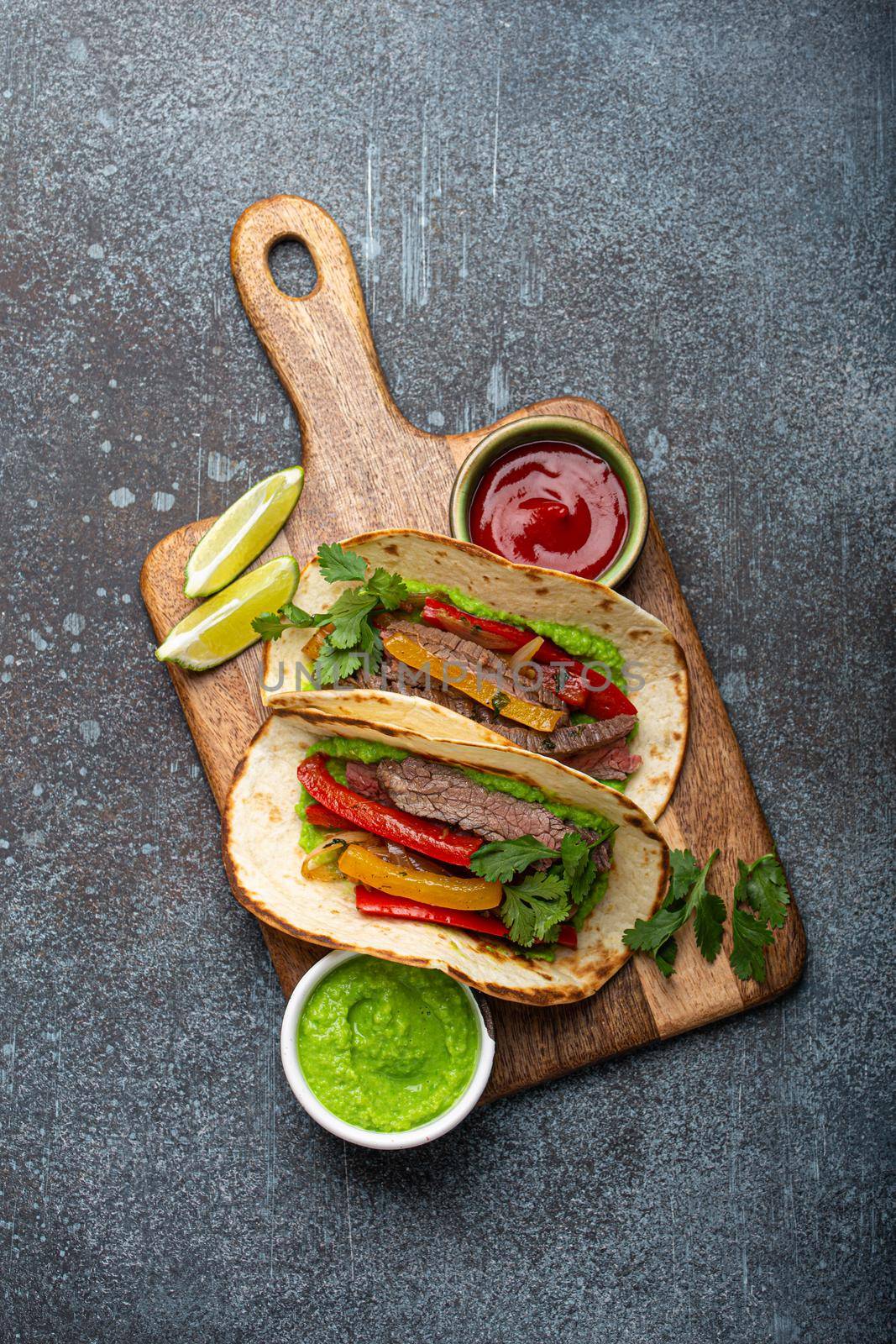 Traditional Mexican dish Beef fajitas tacos served on wooden cutting board with tomato salsa and guacamole on rustic stone background from above, American Mexican food