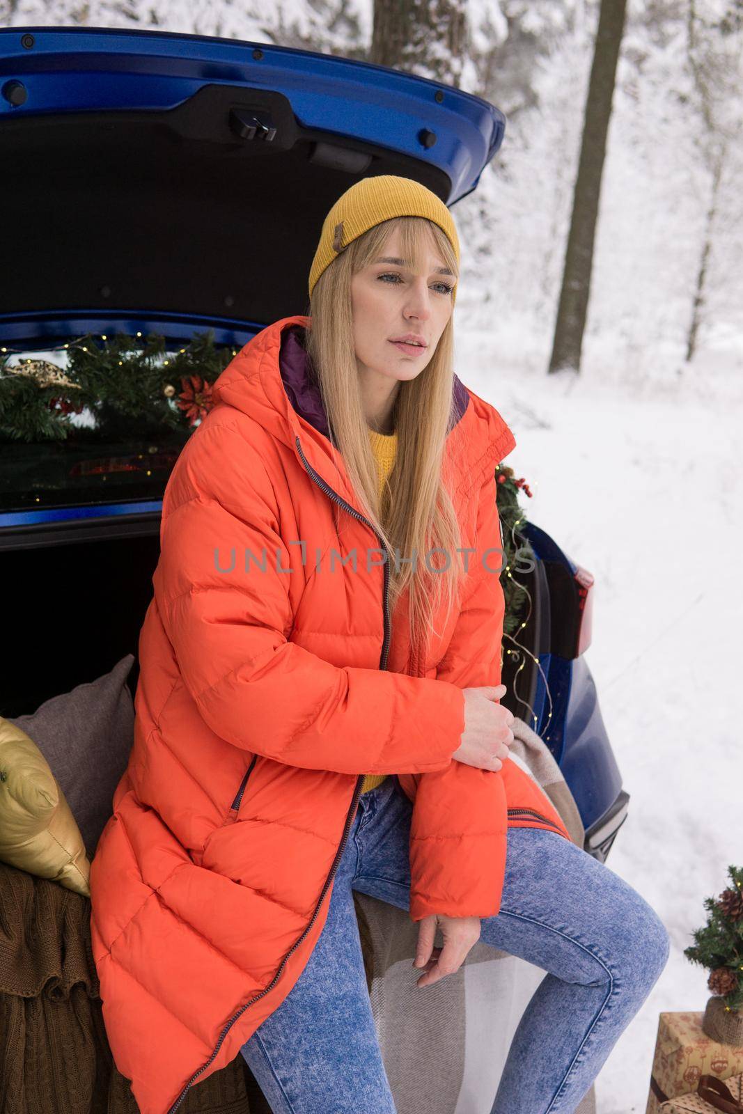 A woman in a winter snow-covered forest in the trunk of a blue car decorated with Christmas decor. The concept of Christmas and winter holidays.