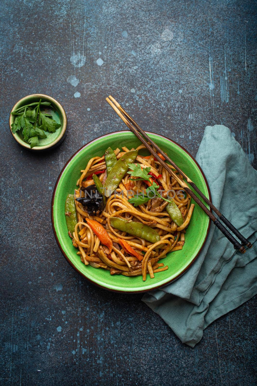 Asian dish stir fry udon noodles with chicken, vegetables and mushrooms in green ceramic bowl with wooden chopsticks on rustic dark blue concrete background from above, Chinese or Thai fast food
