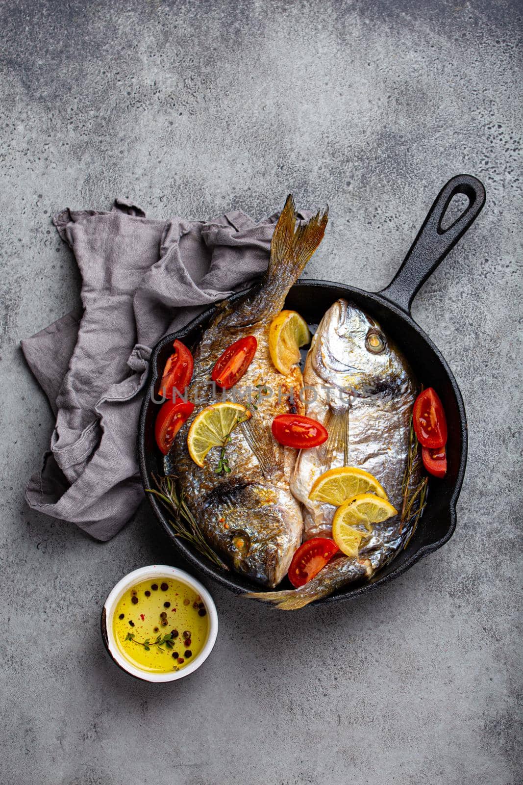 Roasted or fried fresh fish sea bream or dorado with lemon, herbs and tomatoes in cast iron skillet great for healthy meal or Mediterranean diet on rustic grey stone background from above