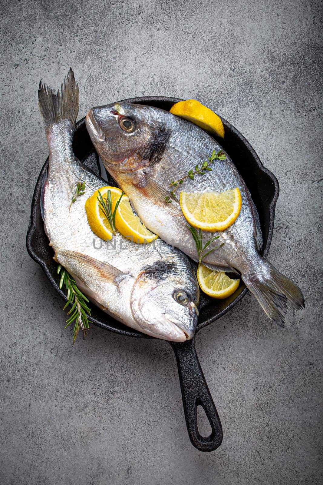 Raw fresh fish sea bream or dorado with lemon, thyme, rosemary and seasonings in cast iron skillet for cooking healthy meal or Mediterranean diet on rustic grey stone background from above