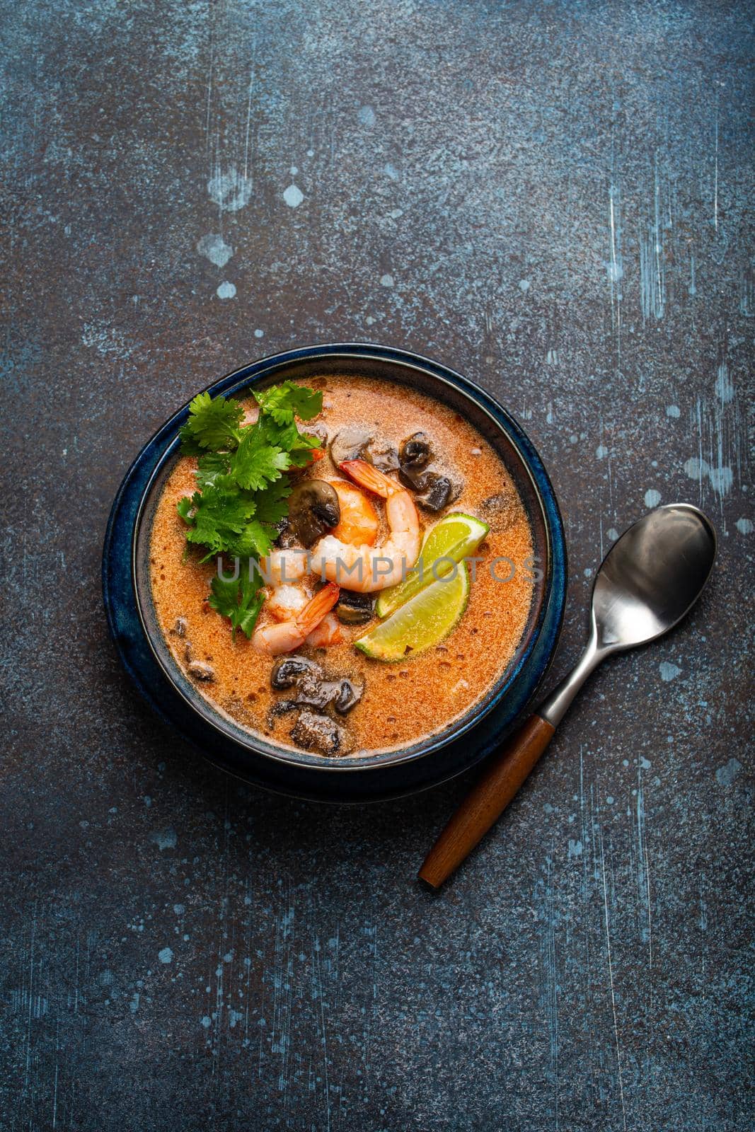 Traditional spicy Thai soup Tom Yum with shrimp and seafood in ceramic bowl on blue stone rustic background from above, classic dish of cuisine of Thailand