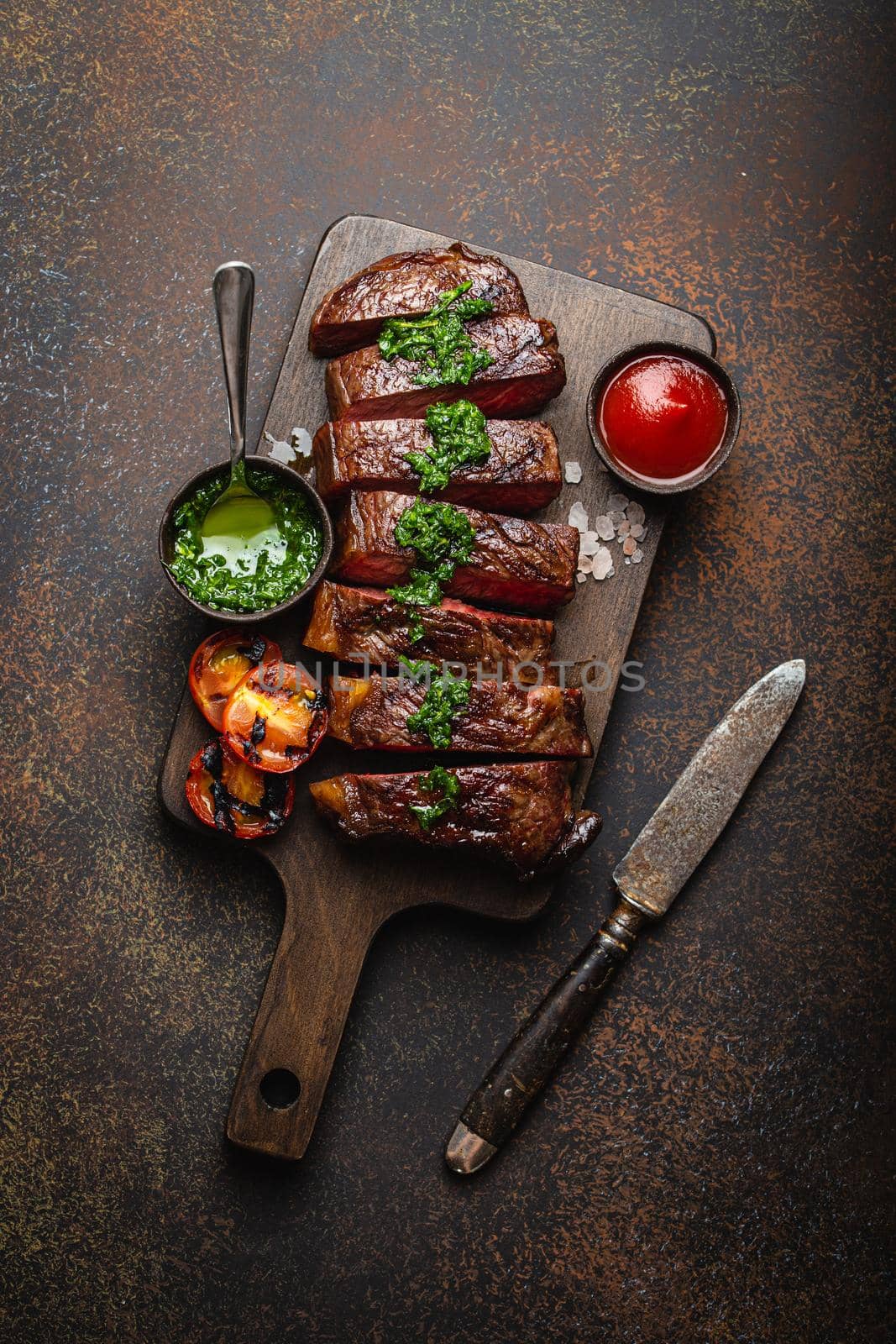 Grilled or fried and sliced marbled meat steak with fork, tomatoes as a side dish and different sauces on wooden cutting board, top view, close-up, stone rustic background. Beef meat steak concept