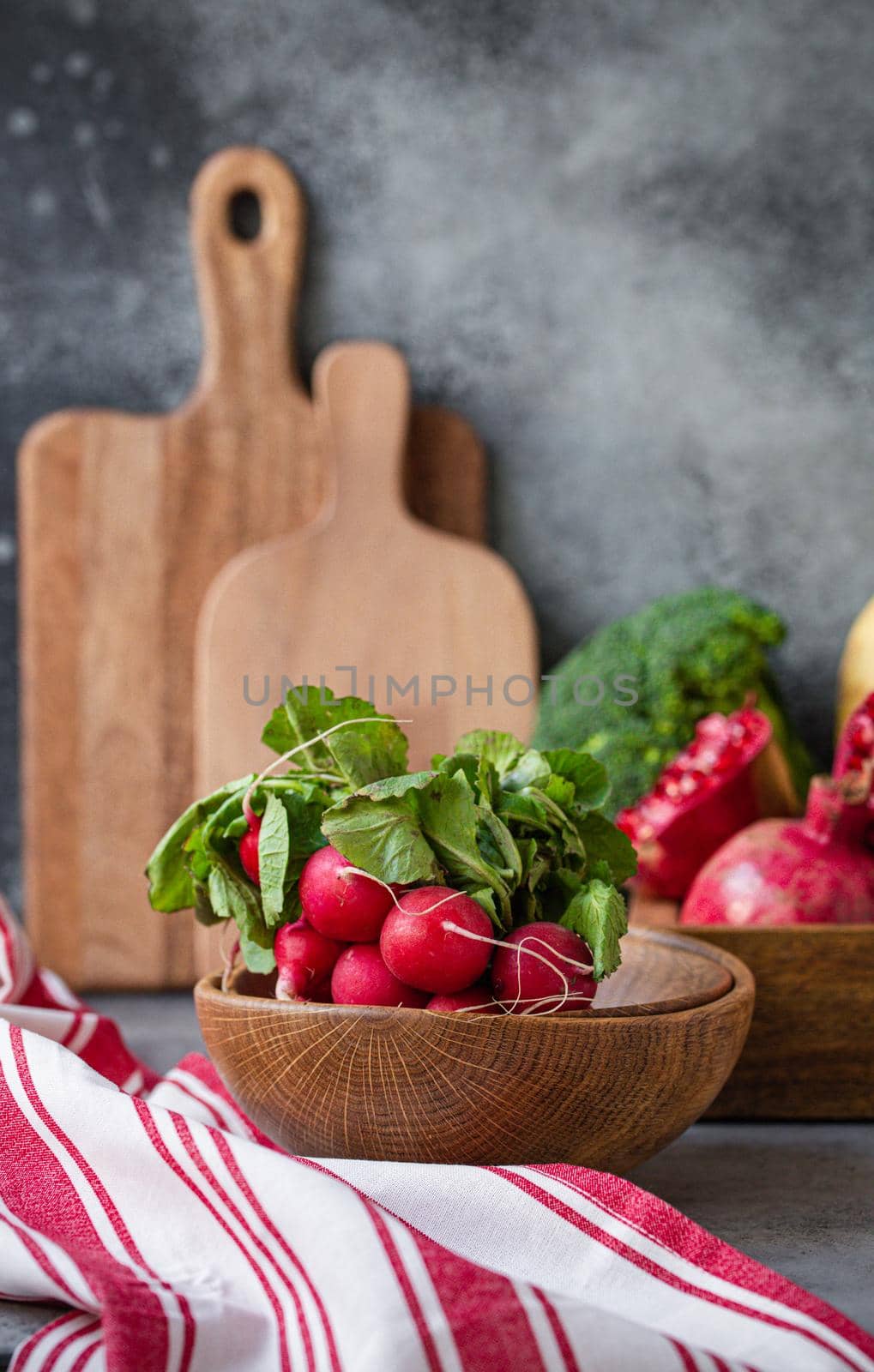Bunch of fresh raw radish in wooden bowl on kitchen table with fresh fruit, greens, vegetables in wooden tray on grey stone background table, cooking healthy diet vegetarian food meal concept.