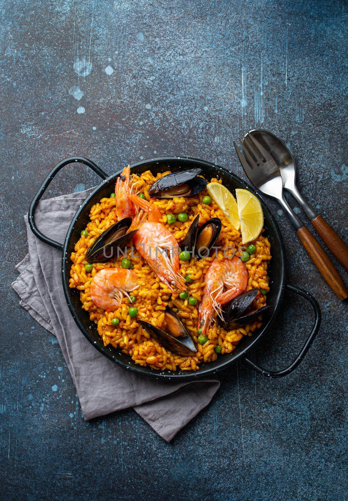 Classic dish of Spain, seafood paella in traditional pan on rustic blue concrete background top view. Spanish paella with shrimps, clamps, mussels, green peas and fresh lemon wedges from above .