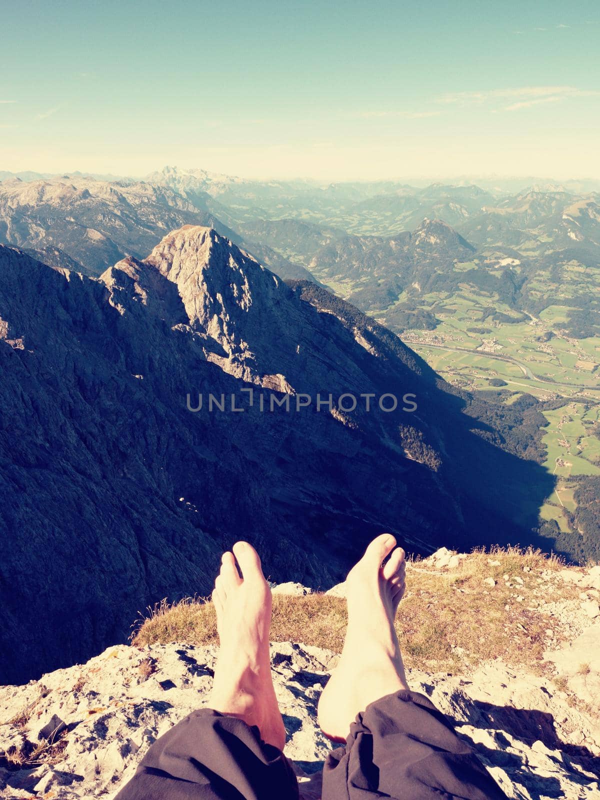 Tired male legs in dark outdoor trousers on peak of mountain above valley. Rest in hike in the mountains. Naked feets without boots on the background of the Alps peaks