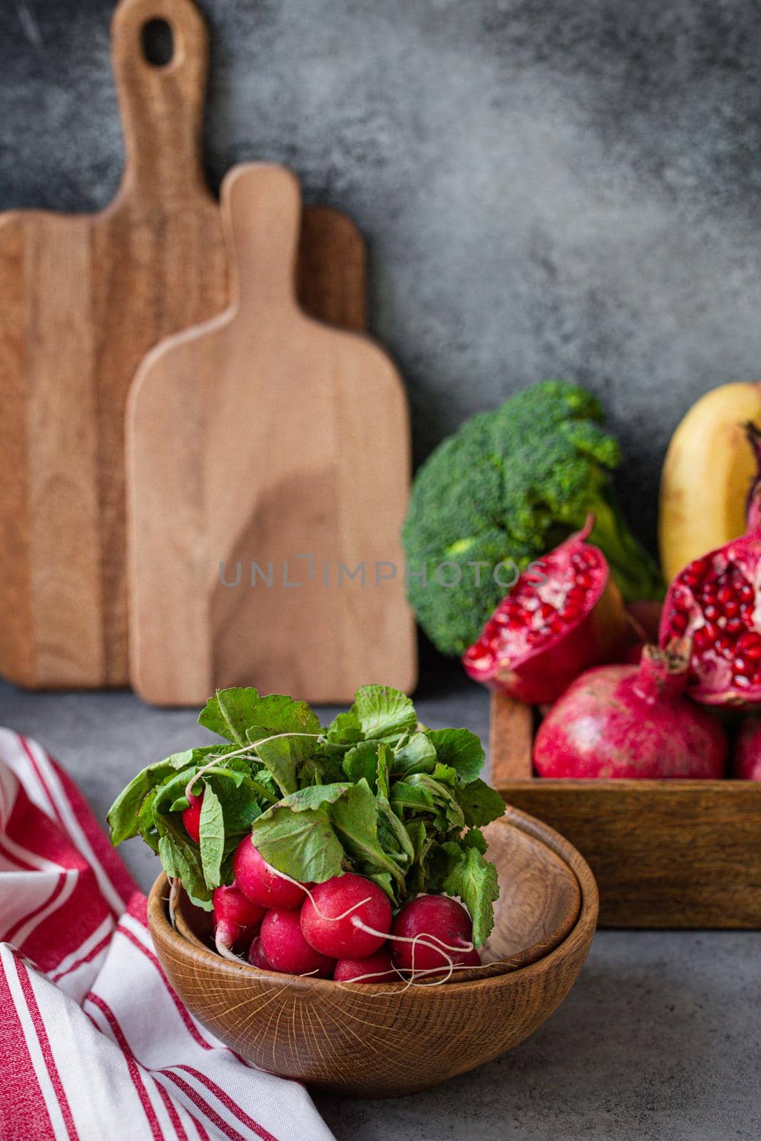 Bunch of fresh raw radish in wooden bowl on kitchen table with fresh fruit, greens, vegetables in wooden tray on grey stone background table, cooking healthy diet vegetarian food meal concept.