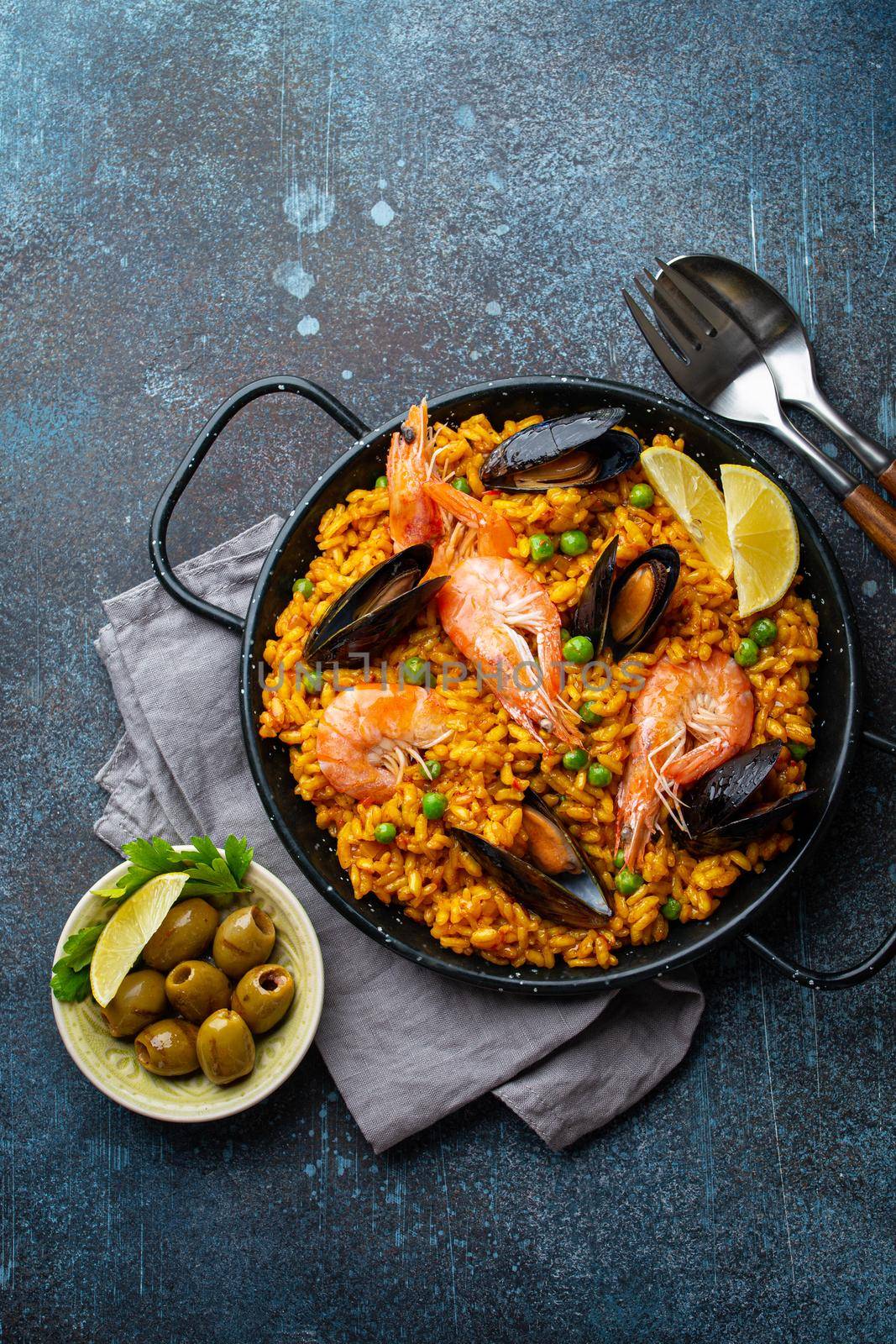 Classic dish of Spain, seafood paella in traditional pan on rustic blue concrete background top view. Spanish paella with shrimps, clamps, mussels, green peas and fresh lemon wedges from above .