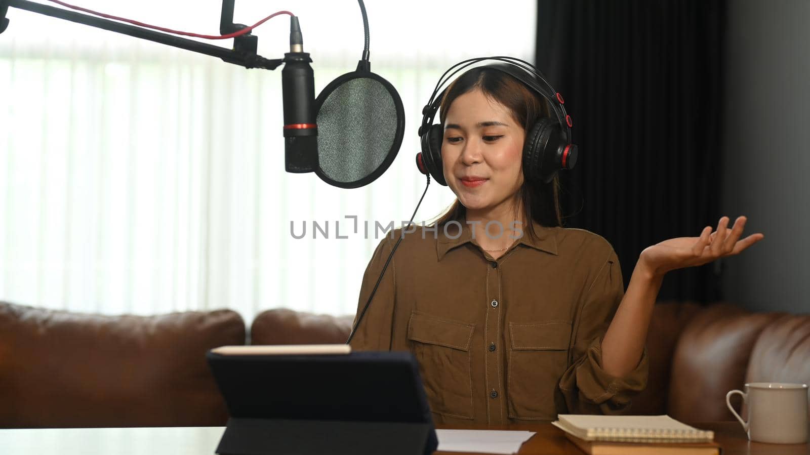 Smiling young woman using laptop and microphone streaming audio podcast at home studio by prathanchorruangsak