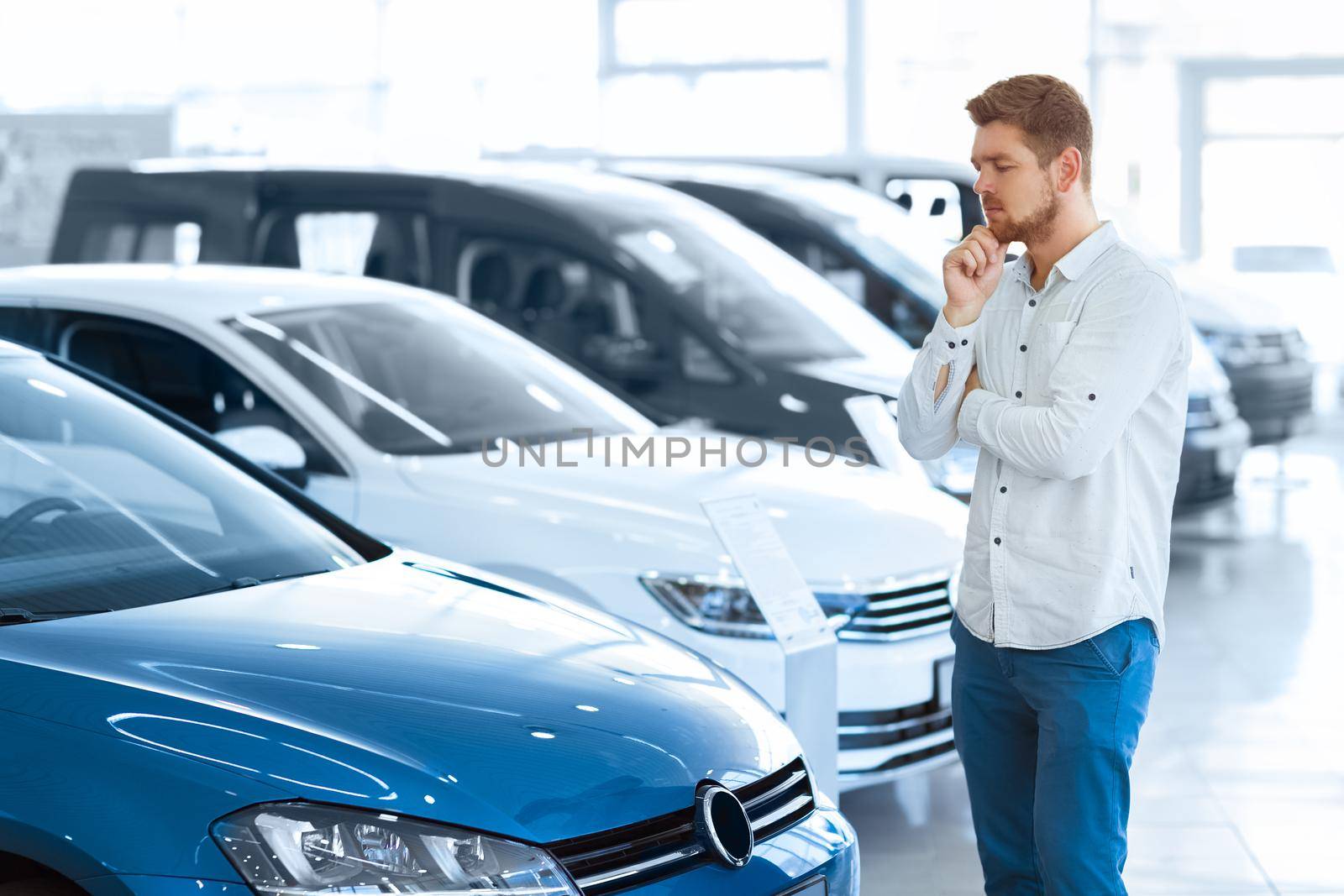 Final decision. Shot of a handsome young man standing in front of a new car at the dealership thinking rubbing his chin